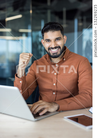 Indian businessman smiling at camera, celebrating achievement with a fist pump while working on a laptop at an office desk, symbolizing success, triumph, and happiness in a corporate environment Indian businessman smiling at camera, celebrating achievement with a fist pump while working on a laptop at an office desk, symbolizing success, triumph, and happiness in a corporate environment 132269235