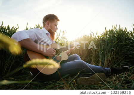 Guitar in hands. Man in white shirt is on the agricultural field Guitar in hands. Man in white shirt is on the agricultural field 132269236
