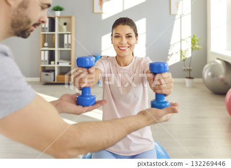 Happy woman doing sport exercise using dumbbells with physiotherapist in clinic. 132269446