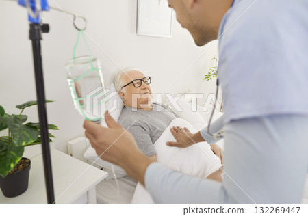 Doctor wearing white coat is placing intravenous line on elderly man lying in hospital bed. 132269447