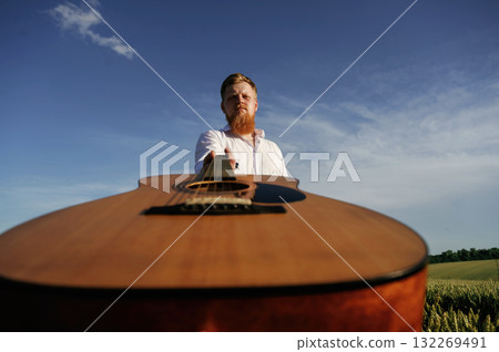Holding acoustic guitar. Man in white shirt is on the agricultural field Holding acoustic guitar. Man in white shirt is on the agricultural field 132269491