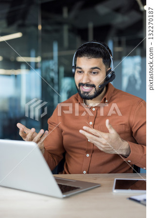 Indian man wearing headset smiling and gesturing hands while having an online video call for customer service support ,on his laptop at a modern office desk Indian man wearing headset smiling and gesturing hands while having an online video call for customer service support ,on his laptop at a modern office desk 132270187