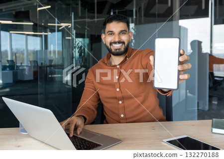 Smiling indian businessman sitting at the office desk with a laptop, holding up a smartphone with an empty white screen for a mock-up advertisement or mobile app presentation 132270188