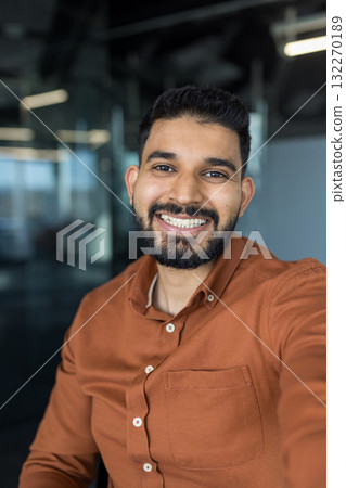 Smiling indian businessman with a beard taking a selfie, showing a happy and positive expression, standing confidently in a modern office or coworking space, representing success and professionalism 132270189