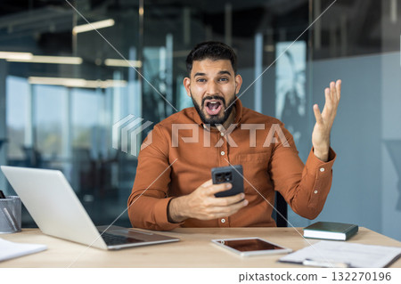 Businessman stares at his smartphone in shock and frustration, mouth agape and hand outstretched as he reacts to bad news at his office desk, stressed and overwhelmed 132270196