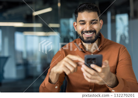Indian businessman smiling, enjoying connection, holding a mobile phone in a modern office, typing message or checking social media, representing communication and technology Indian businessman smiling, enjoying connection, holding a mobile phone in a modern office, typing message or checking social media, representing communication and technology 132270201