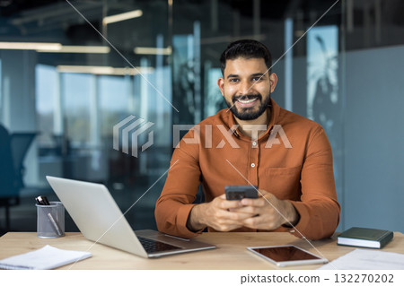 Smiling indian businessman sitting at an office desk, holding a smartphone while working with a laptop and tablet, portraying connectivity and professionalism Smiling indian businessman sitting at an office desk, holding a smartphone while working with a laptop and tablet, portraying connectivity and professionalism 132270202
