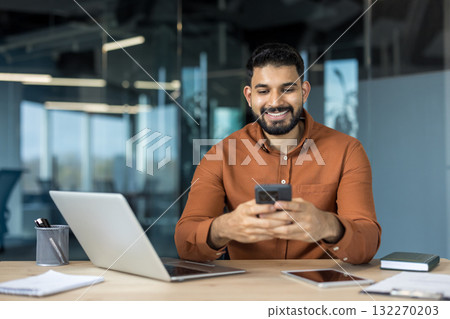 Young indian businessman sitting at his desk in a contemporary office, happily navigating on his smartphone, representing concepts like mobile communication, business technology, and connectivity Young indian businessman sitting at his desk in a contemporary office, happily navigating on his smartphone, representing concepts like mobile communication, business technology, and connectivity 132270203