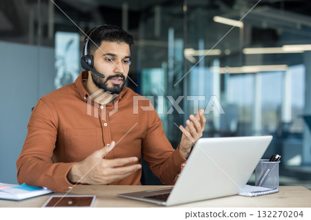 Young bearded man in a brown shirt working in a modern office, having a video conference call on a laptop, actively communicating and gesturing with hands Young bearded man in a brown shirt working in a modern office, having a video conference call on a laptop, actively communicating and gesturing with hands 132270204