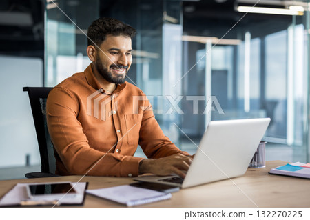 Young indian businessman smiling and typing on a laptop, sitting at a desk in a contemporary office reflecting productivity, success, and professional communication Young indian businessman smiling and typing on a laptop, sitting at a desk in a contemporary office reflecting productivity, success, and professional communication 132270225