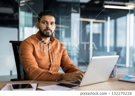 Indian business executive working on a laptop at his desk in a contemporary glass-walled office, focusing on his tasks and representing professionalism and corporate productivity 132270226