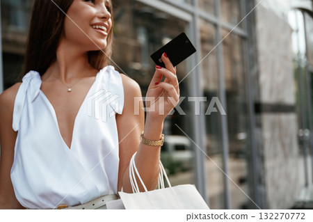 Credit card is in hand. Stylish woman is with shopping bag outdoors near the building Credit card is in hand. Stylish woman is with shopping bag outdoors near the building 132270772