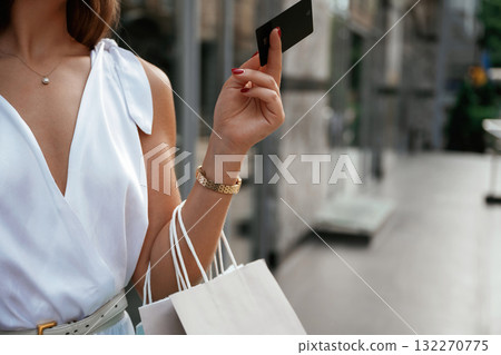 Black credit card in hand. Stylish woman is with shopping bag outdoors near the building Black credit card in hand. Stylish woman is with shopping bag outdoors near the building 132270775