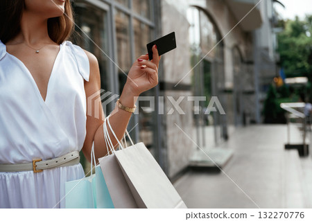 Black credit card in hand. Stylish woman is with shopping bag outdoors near the building Black credit card in hand. Stylish woman is with shopping bag outdoors near the building 132270776