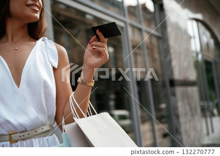 Black credit card in hand. Stylish woman is with shopping bag outdoors near the building 132270777