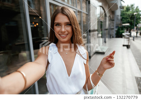 Making a selfie, smiling. Stylish woman is with shopping bag outdoors near the building Making a selfie, smiling. Stylish woman is with shopping bag outdoors near the building 132270779
