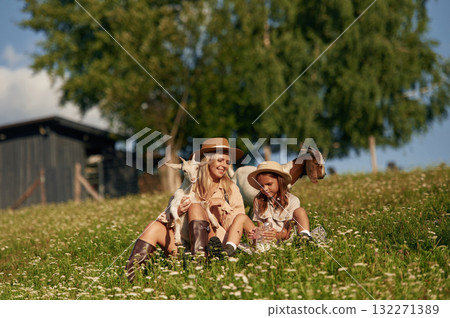 Resting, sitting on the grass. Mother and daughter are together on the farm with goats Resting, sitting on the grass. Mother and daughter are together on the farm with goats 132271389