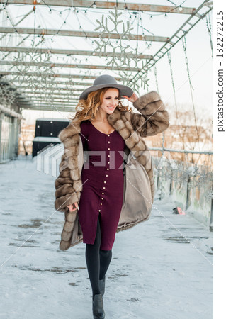 Happy brunette woman standing on snow city street wearing hat and fur coat 132272215