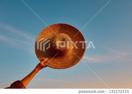 Hand gently lifting straw hat into air against wide blue sky, evoking freedom and summer relaxation, with warm sunlight highlighting woven texture Hand gently lifting straw hat into air against wide blue sky, evoking freedom and summer relaxation, with warm sunlight highlighting woven texture 132272533