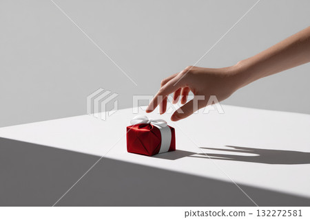 Hand reaching for small red gift box with white ribbon on white table, minimalistic style, soft shadow, elegant and modern composition, joyful and festive mood 132272581