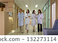 Four female healthcare professionals in white lab coats walking together down modern hospital corridor while smiling and appearing friendly 132273114