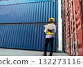 African engineer in yellow hard hat and safety vest holding laptop. Foreman looking to blue shipping container for loading inspection at cargo shipyard. 132273116