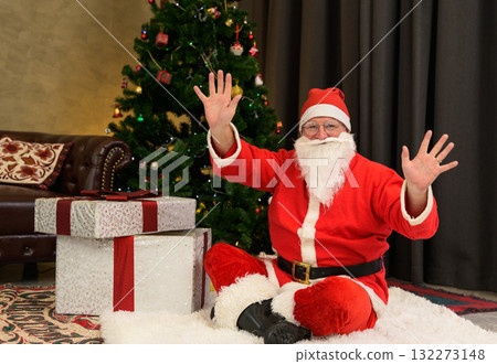 Santa Claus in red suit and hat sitting on white fluffy rug beside decorated Christmas tree while smiling and waving both hands enthusiastically with two large wrapped presents in living room at home 132273148