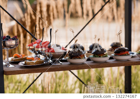 Gourmet Outdoor Catering Spread with Mini Burgers, Fruits, and Appetizers on Wooden Shelf, Rustic Event Setup, Warm Golden Light. Gourmet Outdoor Catering Spread with Mini Burgers, Fruits, and Appetizers on Wooden Shelf, Rustic Event Setup, Warm Golden Light. 132274167