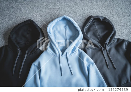 Black, gray, and blue hoodies neatly arranged on smooth gray surface. Casual fashion displayed in natural lighting. Neural network AI generated 132274389
