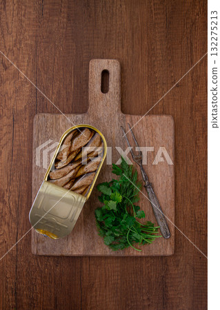 an open can of sprats, appetizer, on a wooden cutting board, with a fork and herbs, top view, an open can of sprats, appetizer, on a wooden cutting board, with a fork and herbs, top view, 132275213
