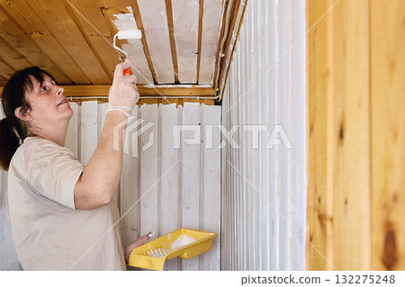 Painting ceiling, woman applying white paint on wooden planks to brighten small interior space and prepare surface for cozy home design. Painting ceiling, woman applying white paint on wooden planks to brighten small interior space and prepare surface for cozy home design. 132275248