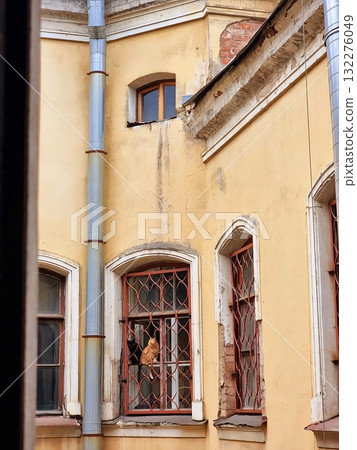 An orange tabby cat sitting on a windowsill behind a rusty grate in a classic St. Petersburg courtyard building. An orange tabby cat sitting on a windowsill behind a rusty grate in a classic St. Petersburg courtyard building. 132276049