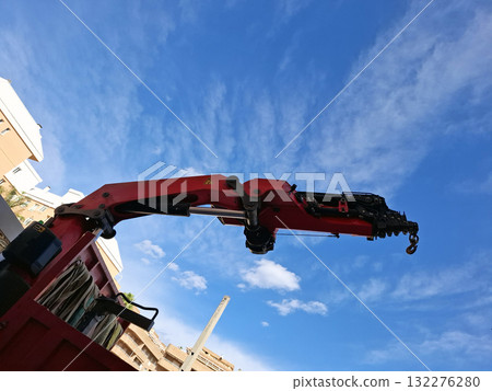 A truck mounted crane extends its telescopic boom high against a clear blue sky filled with clouds. The scene is set in an urban area, showcasing construction activity nearby. A truck mounted crane extends its telescopic boom high against a clear blue sky filled with clouds. The scene is set in an urban area, showcasing construction activity nearby. 132276280