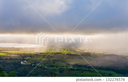 A sweeping valley landscape with green fields, winding roads, and misty sunrays 132276576
