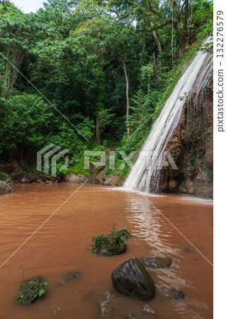 A serene tropical scene featuring a tall waterfall pouring over a rocky cliff A serene tropical scene featuring a tall waterfall pouring over a rocky cliff 132276579
