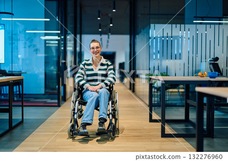 A confident smiling business woman in a wheelchair sitting in a bright modern office hallway, representing workplace inclusion, accessibility, and professional success. 132276960