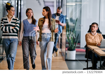 Group of coworkers walking and discussing ideas in a modern office corridor while a male colleague works at a laptop nearby, symbolizing teamwork, productivity, and creative collaboration in a 132276984