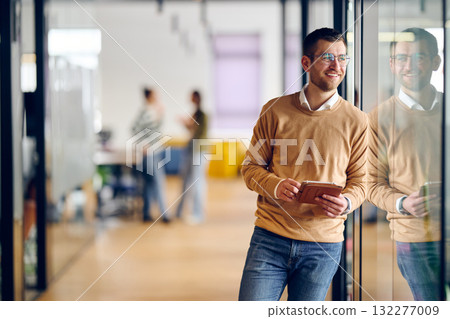 Businessman standing by glass wall in a modern office corridor, holding a digital tablet and smiling with reflection visible on the glass, symbolizing success, optimism, and modern business lifestyle. 132277009