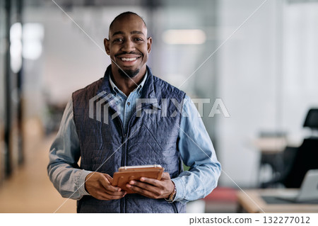 Confident african american businessman standing in a modern office hallway, smiling while using a digital tablet. Reflection in glass wall symbolizes technology, success, and innovation in business. 132277012