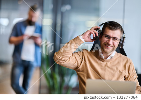 Happy businessman sitting at office desk, wearing headphones and relaxing while listening to music during a break, representing stress relief, balance, and positive work atmosphere in modern office Happy businessman sitting at office desk, wearing headphones and relaxing while listening to music during a break, representing stress relief, balance, and positive work atmosphere in modern office 132277064