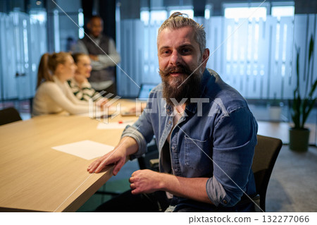 Portrait of a smiling confident businessman with beard sitting at conference table during a team meeting in a modern office, symbolizing leadership, collaboration, and teamwork 132277066