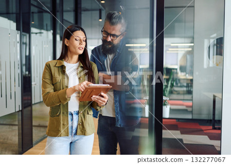Two business professionals having a focused discussion while reviewing information on a tablet in a bright modern office hallway, symbolizing teamwork, communication, and digital collaboration. Two business professionals having a focused discussion while reviewing information on a tablet in a bright modern office hallway, symbolizing teamwork, communication, and digital collaboration. 132277067