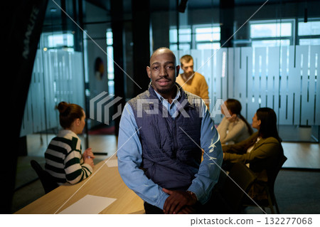 Portrait of a confident african american businessman in a modern office with colleagues discussing in the background, symbolizing leadership, teamwork, success, and collaboration in a corporate 132277068