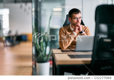 Focused businessman sitting at desk with laptop, thoughtfully analyzing data or planning strategy in a modern office environment, representing concentration, leadership, and innovation. 132277069