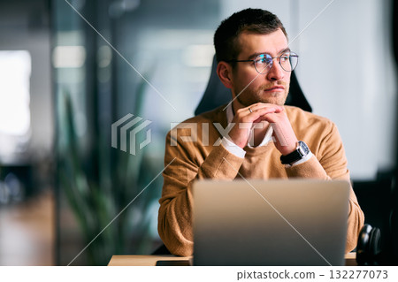 Focused businessman sitting at desk with laptop, thoughtfully analyzing data or planning strategy in a modern office environment, representing concentration, leadership, and innovation. 132277073