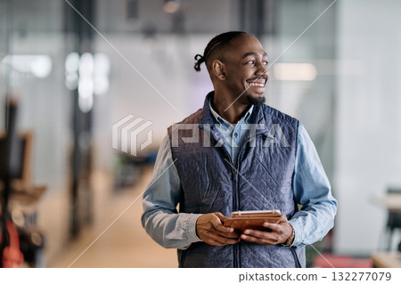Confident african american businessman standing in a modern office hallway, smiling while using a digital tablet. Reflection in glass wall symbolizes technology, success, and innovation in business. Confident african american businessman standing in a modern office hallway, smiling while using a digital tablet. Reflection in glass wall symbolizes technology, success, and innovation in business. 132277079