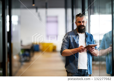 Bearded businessman standing in modern office hallway, holding digital tablet and looking confidently at camera, representing experience, leadership, and innovation in contemporary business 132277082