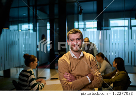 Portrait of a smiling young businessman standing confidently in a modern office with his team collaborating in the background, representing leadership, teamwork, confidence, and corporate success. 132277088
