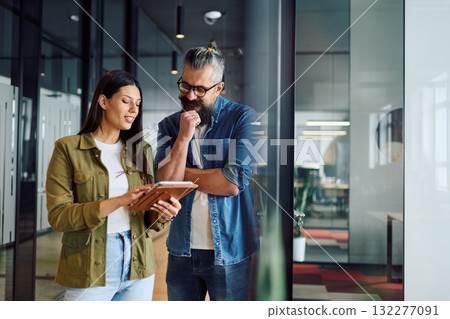 Two business professionals having a focused discussion while reviewing information on a tablet in a bright modern office hallway, symbolizing teamwork, communication, and digital collaboration. 132277091