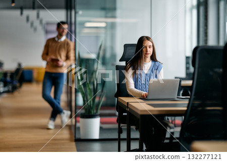 Businesswoman sitting at desk and working on laptop in a bright modern office while colleague stands in the background, representing productivity, focus, and teamwork in a professional environment. 132277121